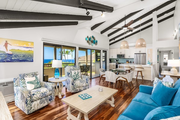 A spacious living area with a wooden floor, a blue sofa, patterned armchairs, a dining table, and a kitchen in the background.