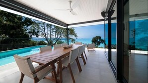 A covered outdoor dining area with a view of the sea, a pool, and a ceiling fan.