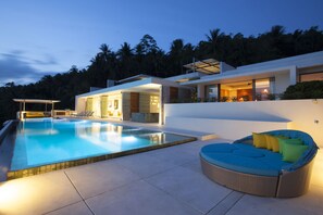 Une maison moderne dotée d’une piscine, d’un patio couvert et d’un espace détente avec des coussins.