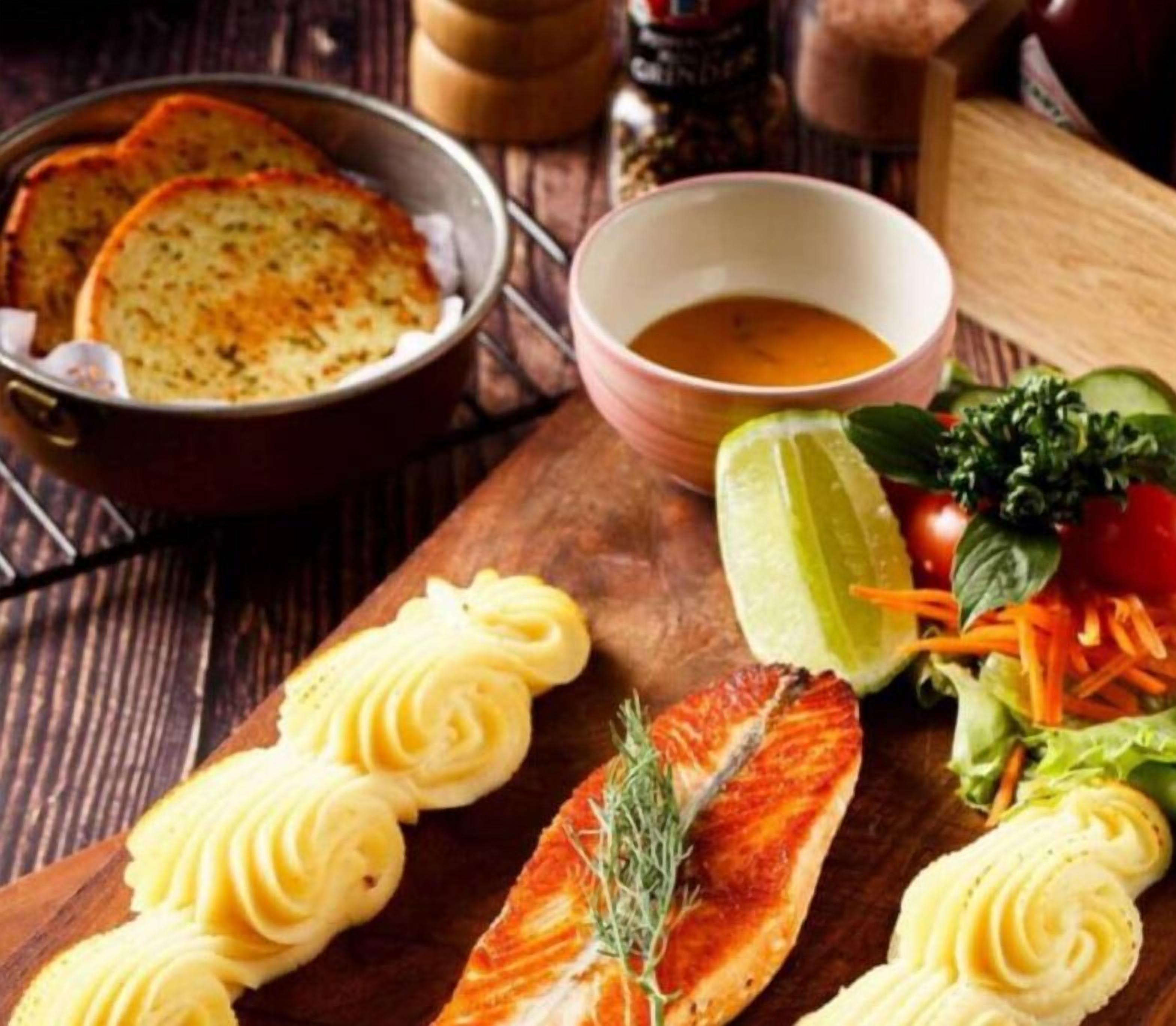 Une table en bois sur laquelle on trouve un filet de saumon, un bol de soupe et une assiette de pain tranché.