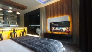 Habitació d'hotel amb un llit, una zona de bar amb tamborets, una televisió penjada a la paret i un gran finestral.