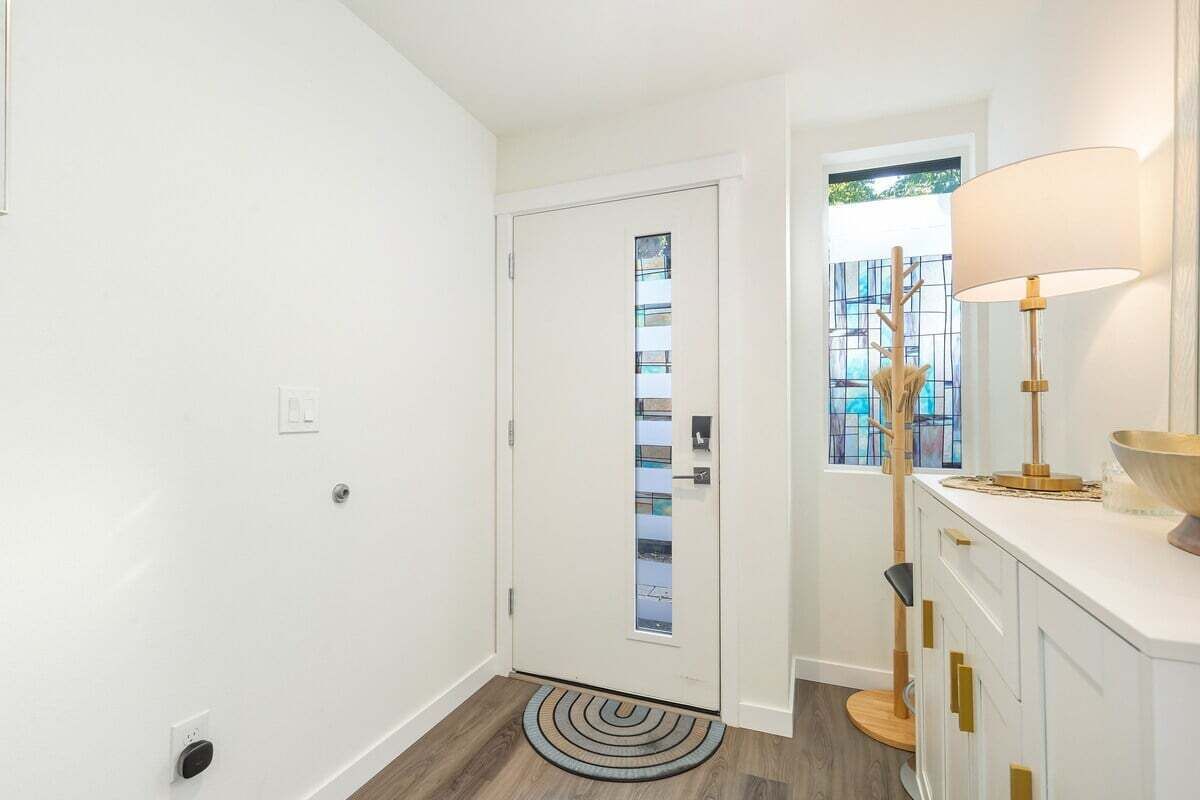 A hallway with a white door, a wooden floor, a patterned rug, a modern lamp, and a window with a view of greenery.