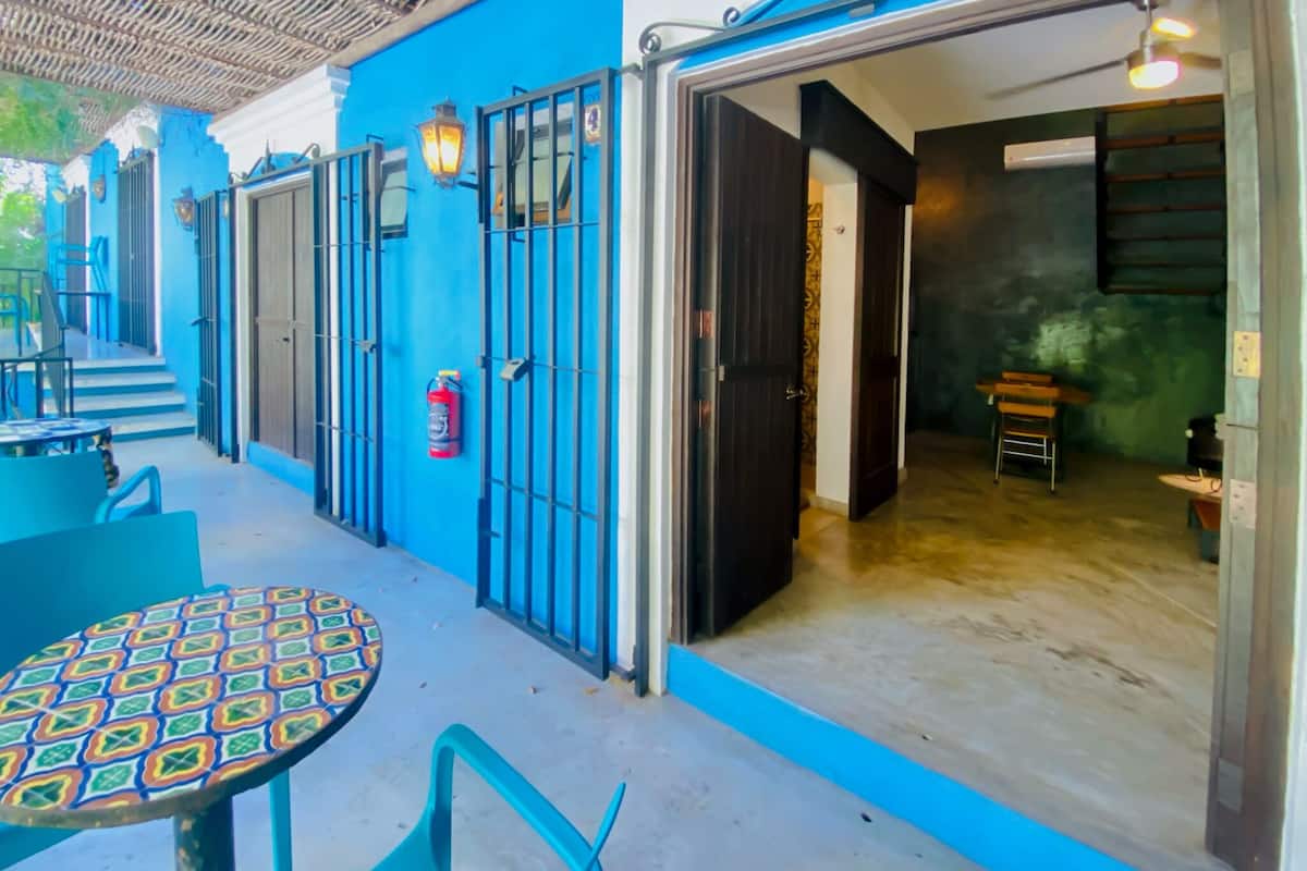Une façade bleue, une entrée fermée par un portail, un extincteur et une table avec des chaises à l’extérieur.