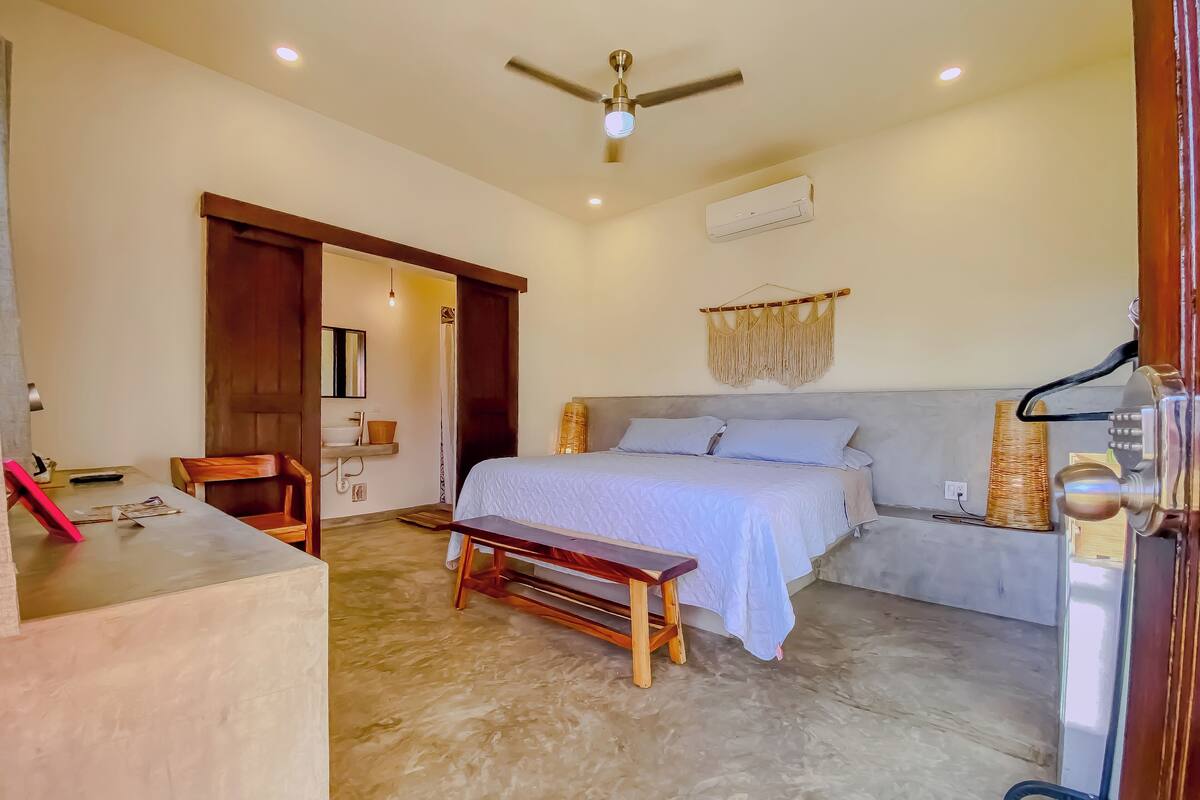 Une chambre à coucher comprenant un lit, une table de chevet, un banc, un ventilateur de plafond et un climatiseur fixé au mur.