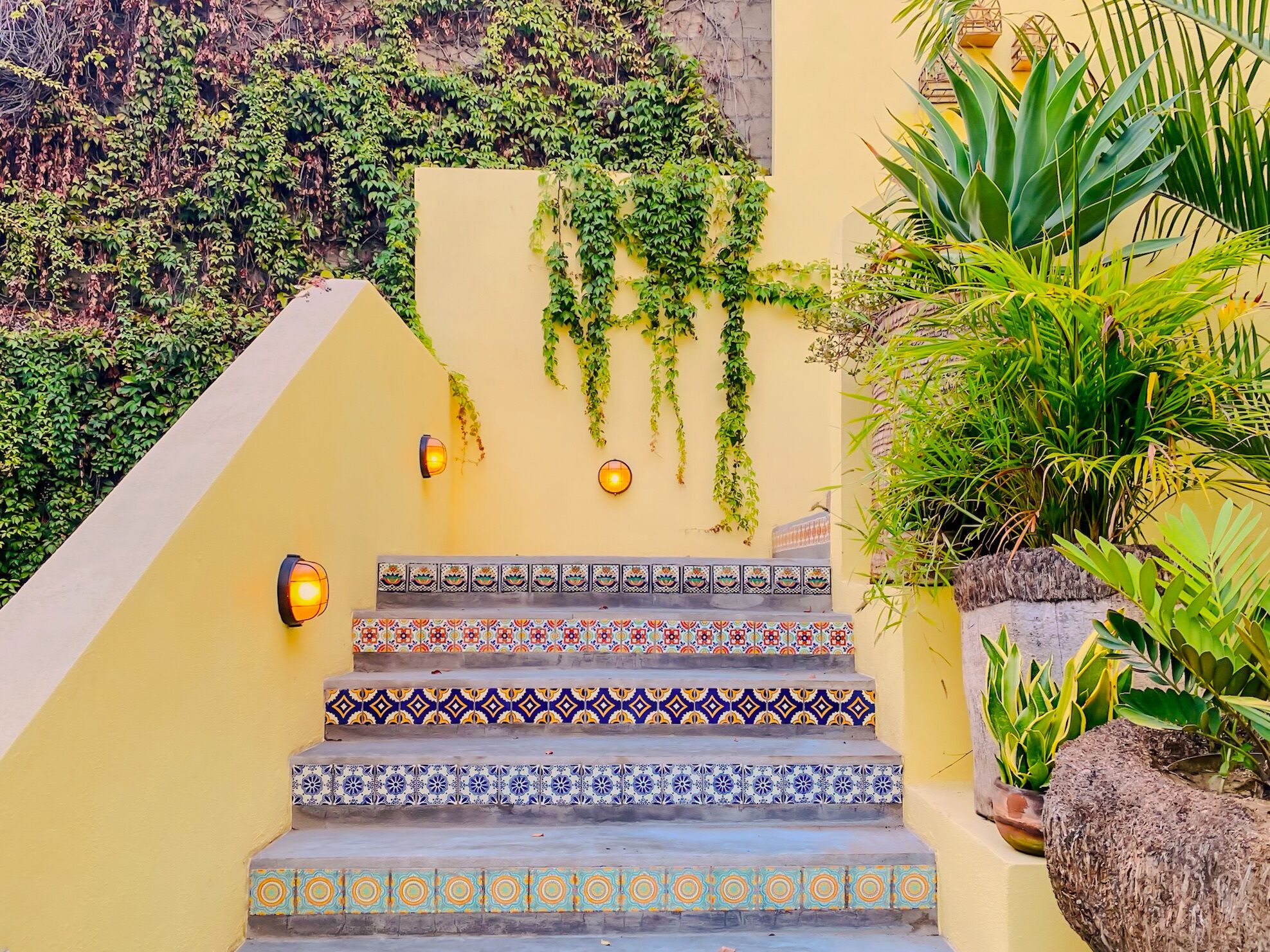 A set of stairs with colorful patterned tiles, surrounded by lush greenery and decorative wall lights.