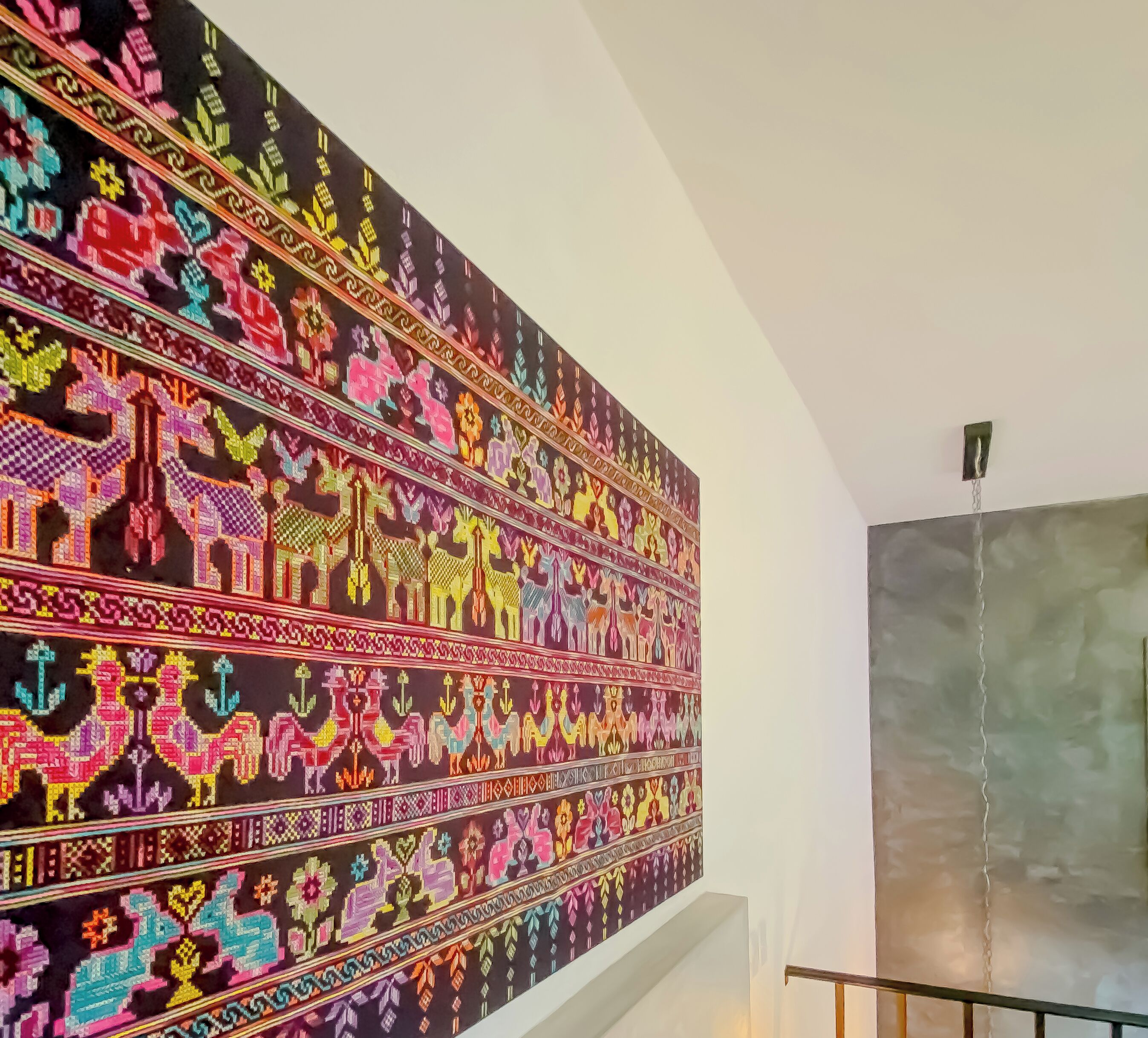 Ein großes, farbenfrohes Wandteppich mit komplexen Mustern und Figuren, präsentiert an einer Wand in einem Zimmer mit schlichtem, modernem Design.