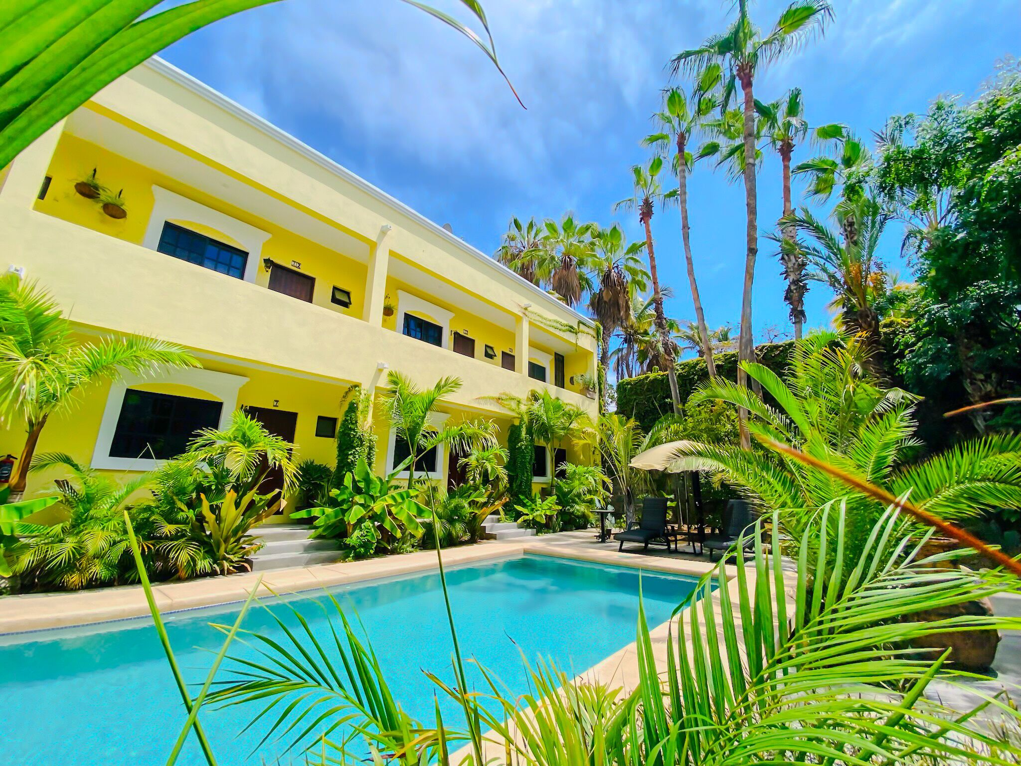 A two-story yellow building with a swimming pool surrounded by tropical plants.