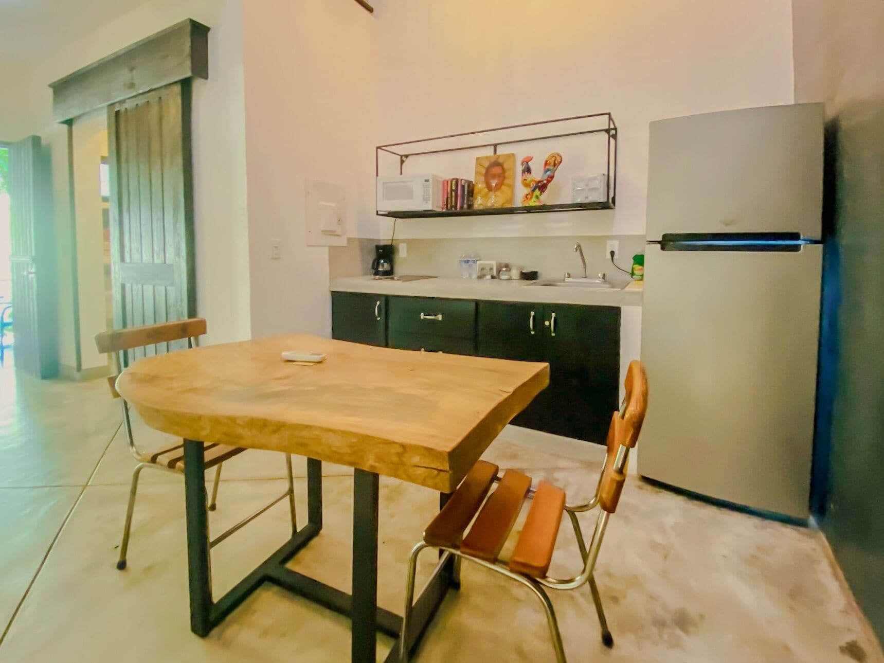 A small kitchen with a wooden table and two chairs, a microwave shelf, and a refrigerator.