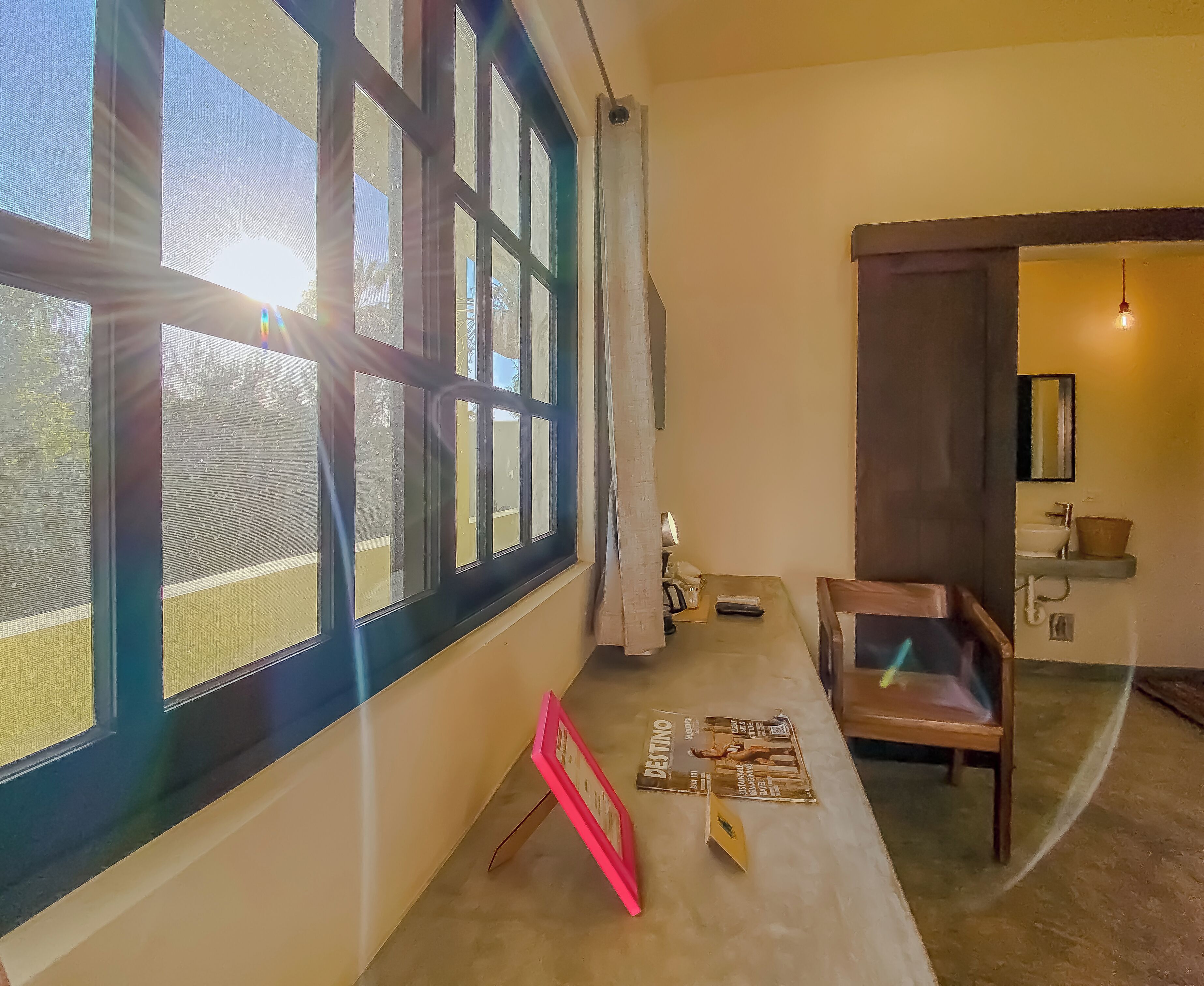 A room with large windows, a wooden chair, a table with magazines, and a bathroom with a sink and mirror.