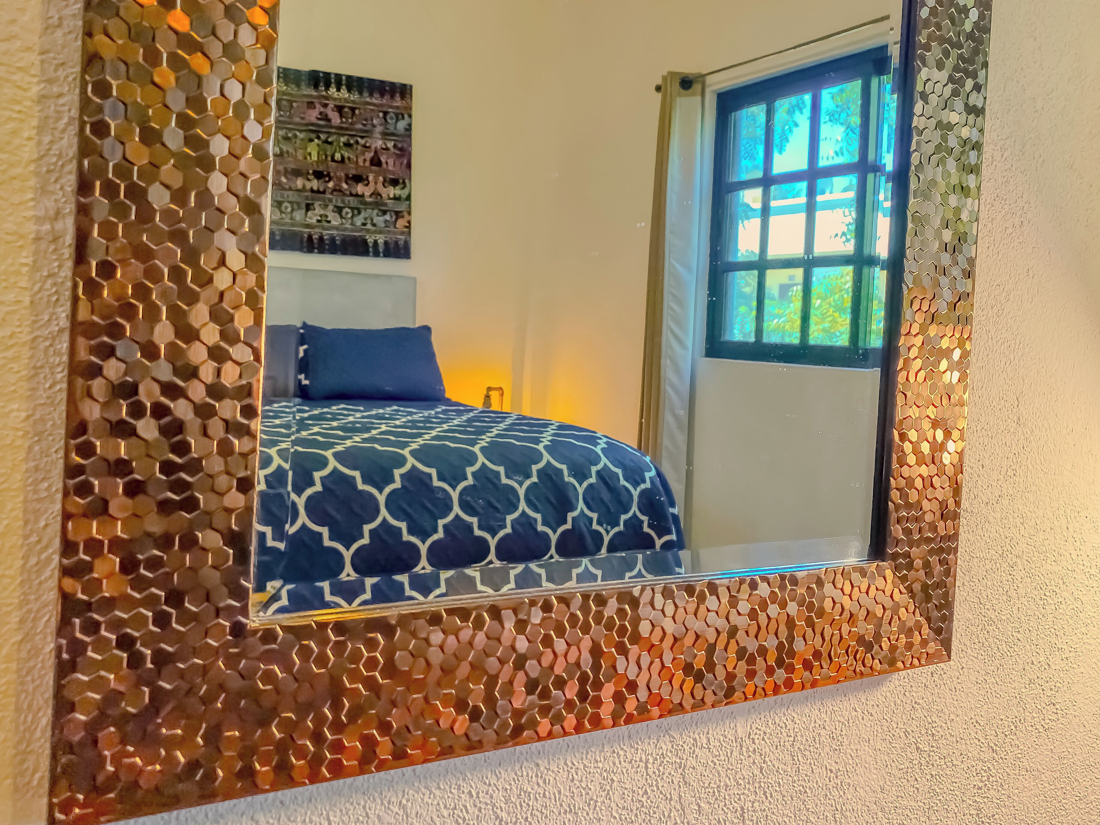 Un dormitorio con un espejo grande, una cama con colcha estampada y una ventana que da a zonas verdes.