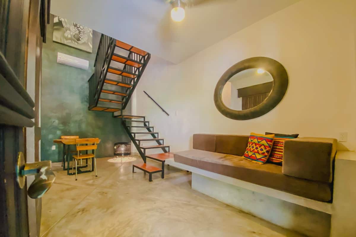 Un salon moderne avec un escalier, un grand miroir rond, un canapé agrémenté de coussins colorés et une petite table avec des chaises.