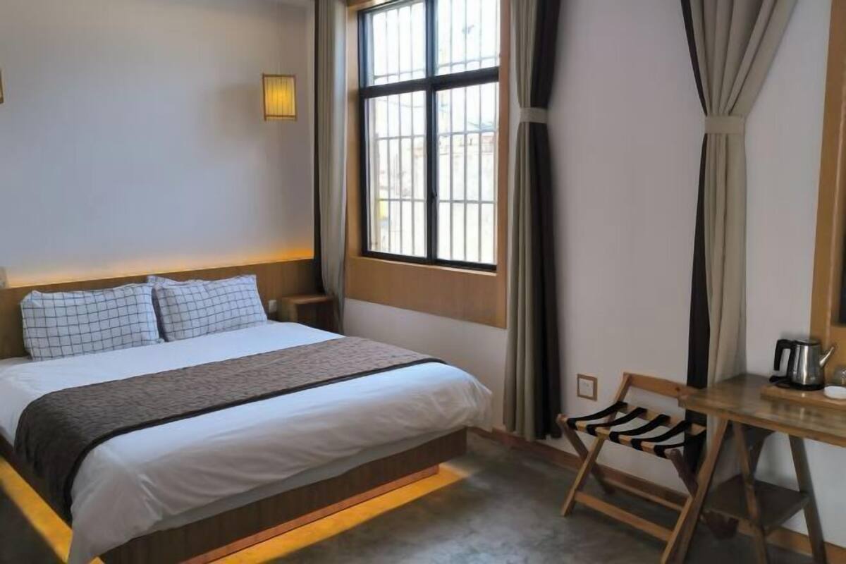 A hotel room with a bed, a desk, a chair, a window with curtains, and a lamp.
