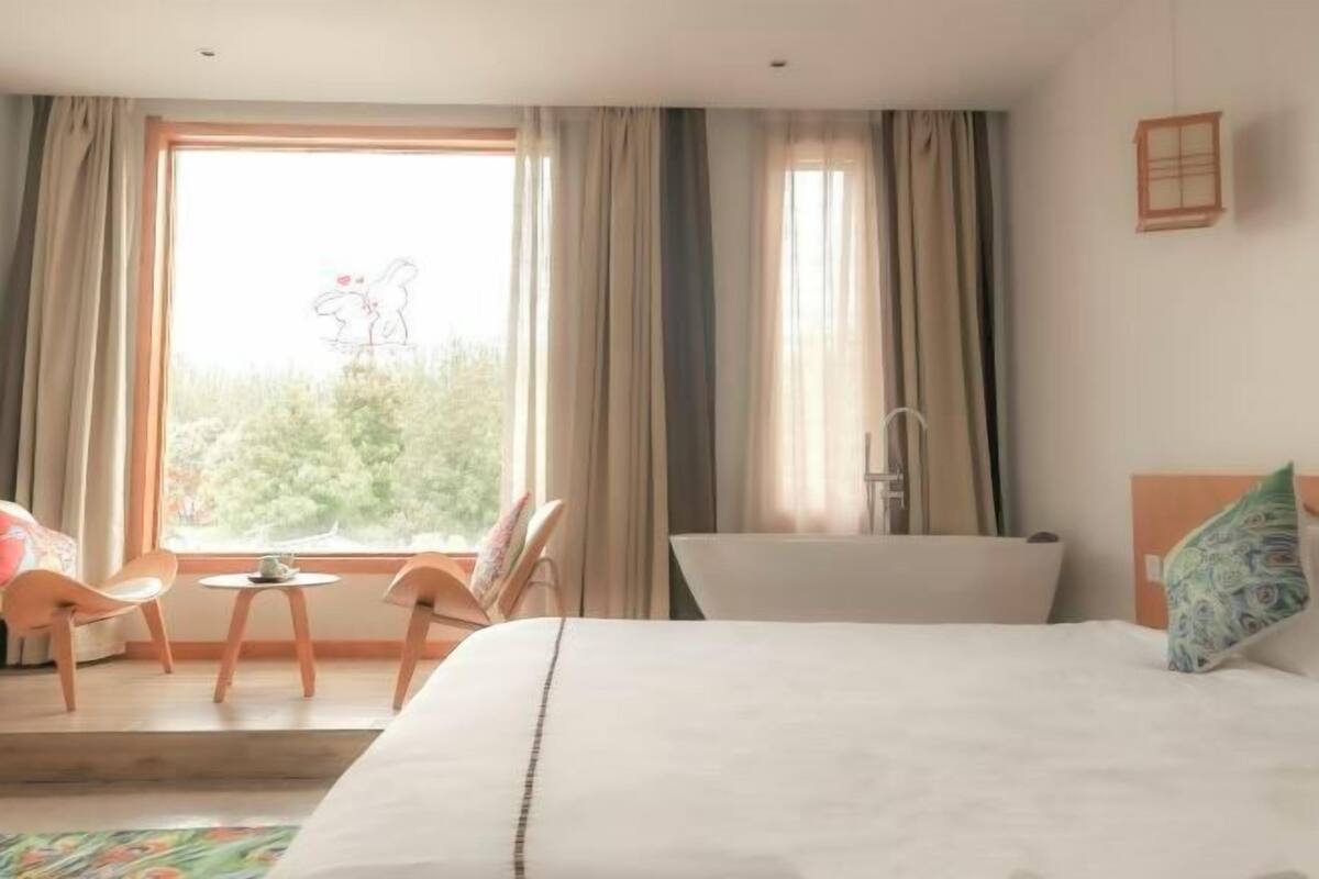 A bedroom with a bed, a window with curtains, a small table, two chairs, and a bathtub.
