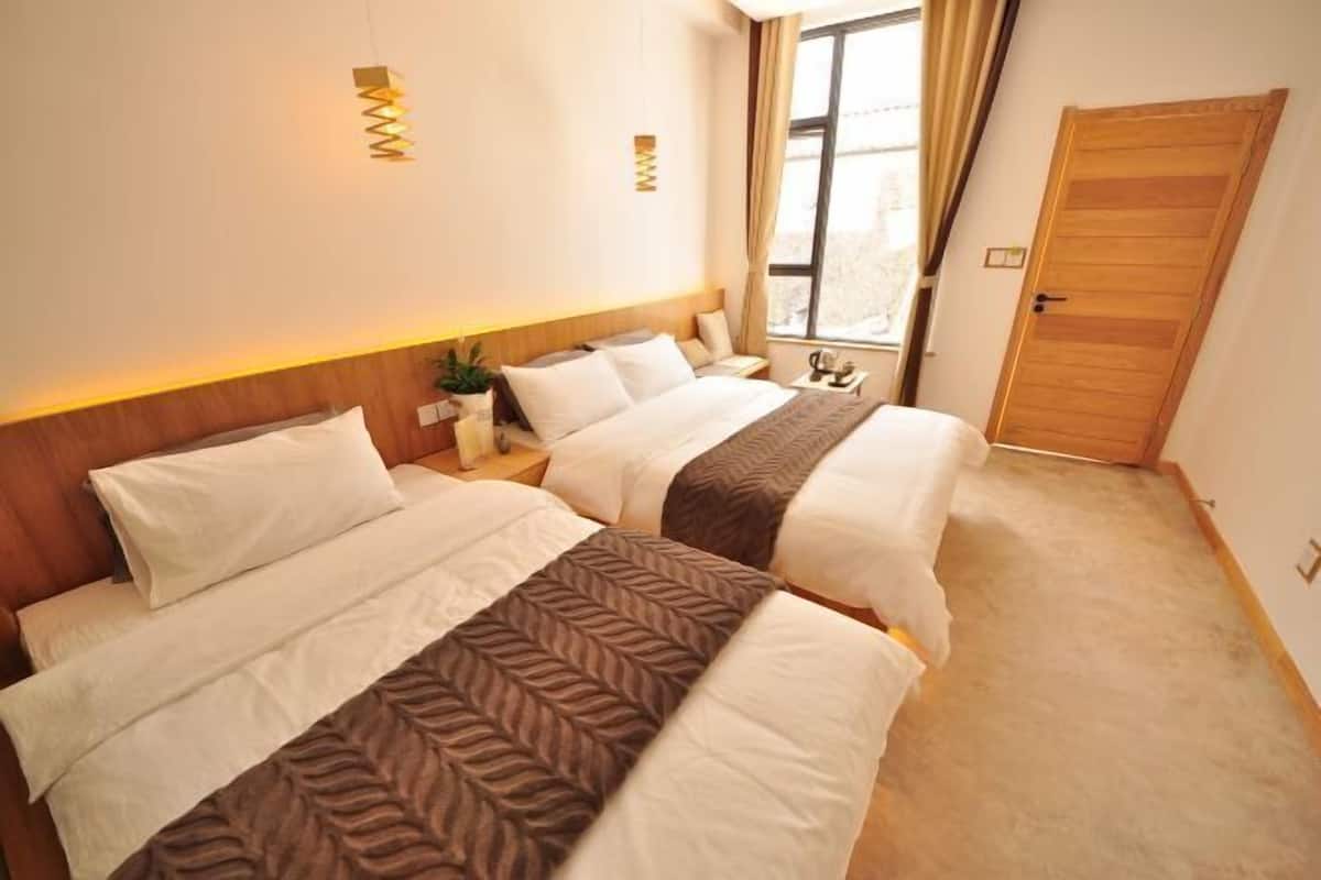 A hotel room with two beds, a wooden headboard, a small bedside table with a lamp, and a window with curtains.