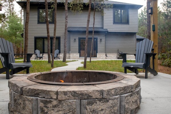 Una casa moderna con un hogar de piedra y sillas Adirondack en el jardín delantero.