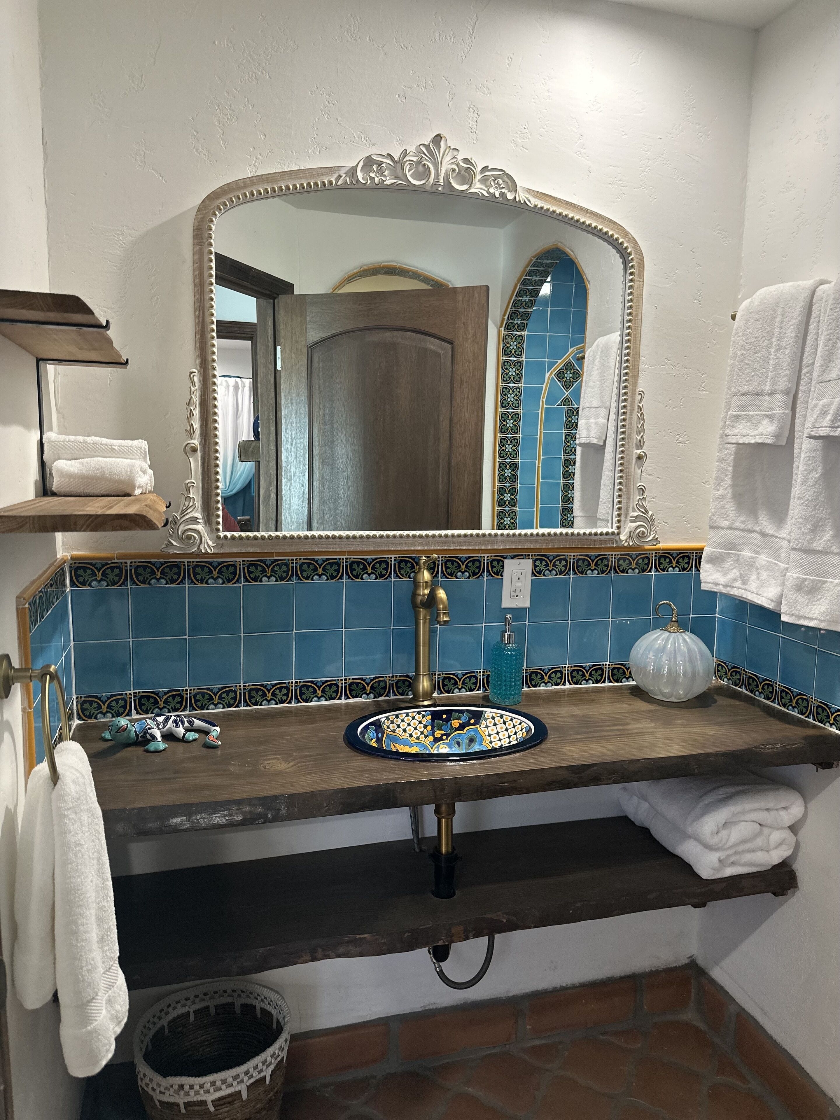 Une salle de bain avec un meuble-lavabo en bois, un grand miroir, un pare-chocs bleu en carreaux, un lavabo blanc et une poubelle blanche.