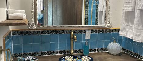 Une salle de bain avec un meuble-lavabo en bois, un grand miroir, un pare-chocs bleu en carreaux, un lavabo blanc et une poubelle blanche.