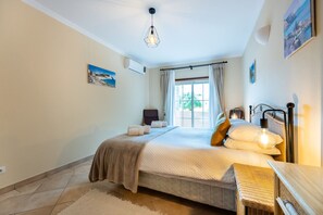 Un dormitorio con cama, una mesita de noche, lámpara, silla, ventana con cortinas y aire acondicionado instalado en la pared.