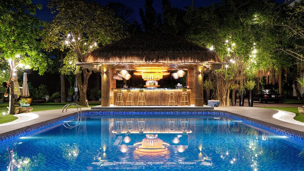 A poolside bar area with a thatched roof, bar stools, and a pool with blue tiles.