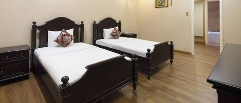 Une chambre d’hôtel avec deux lits en bois, une table de chevet, une commode et une salle de bain visible à travers une porte ouverte.