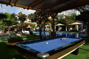 Une table de billard avec des billes, entourée de sièges et de parasols, avec une piscine en arrière-plan.