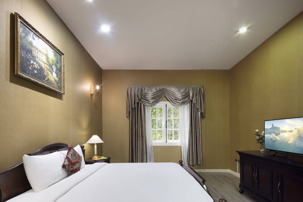 Een hotelkamer met een groot bed, een televisie, een nachtkastje, een lamp, een schilderij aan de muur en een raam met gordijnen.
