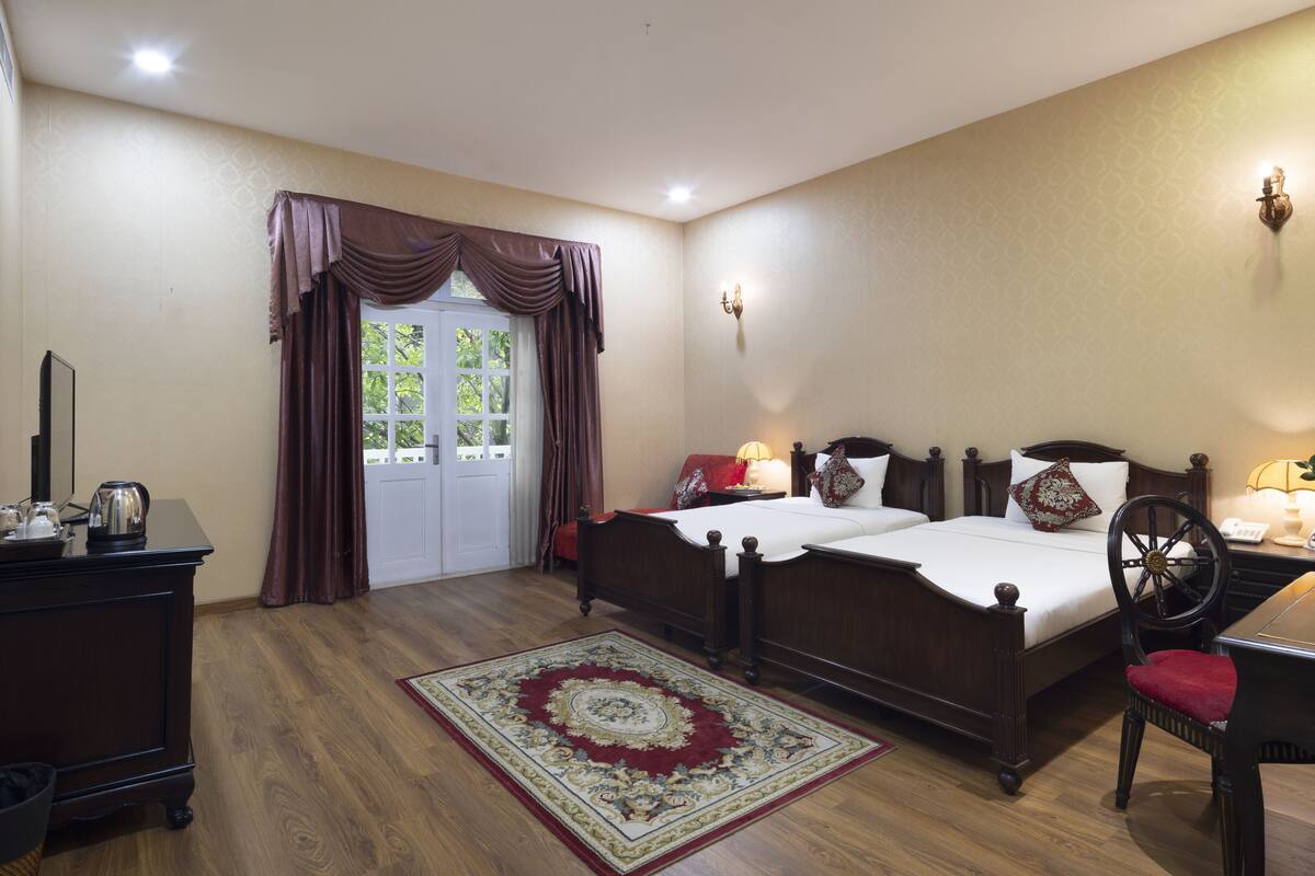 Een slaapkamer met twee bedden, een houten dressoir, een bureau met stoel, een vloerkleed en een raam met gordijnen.