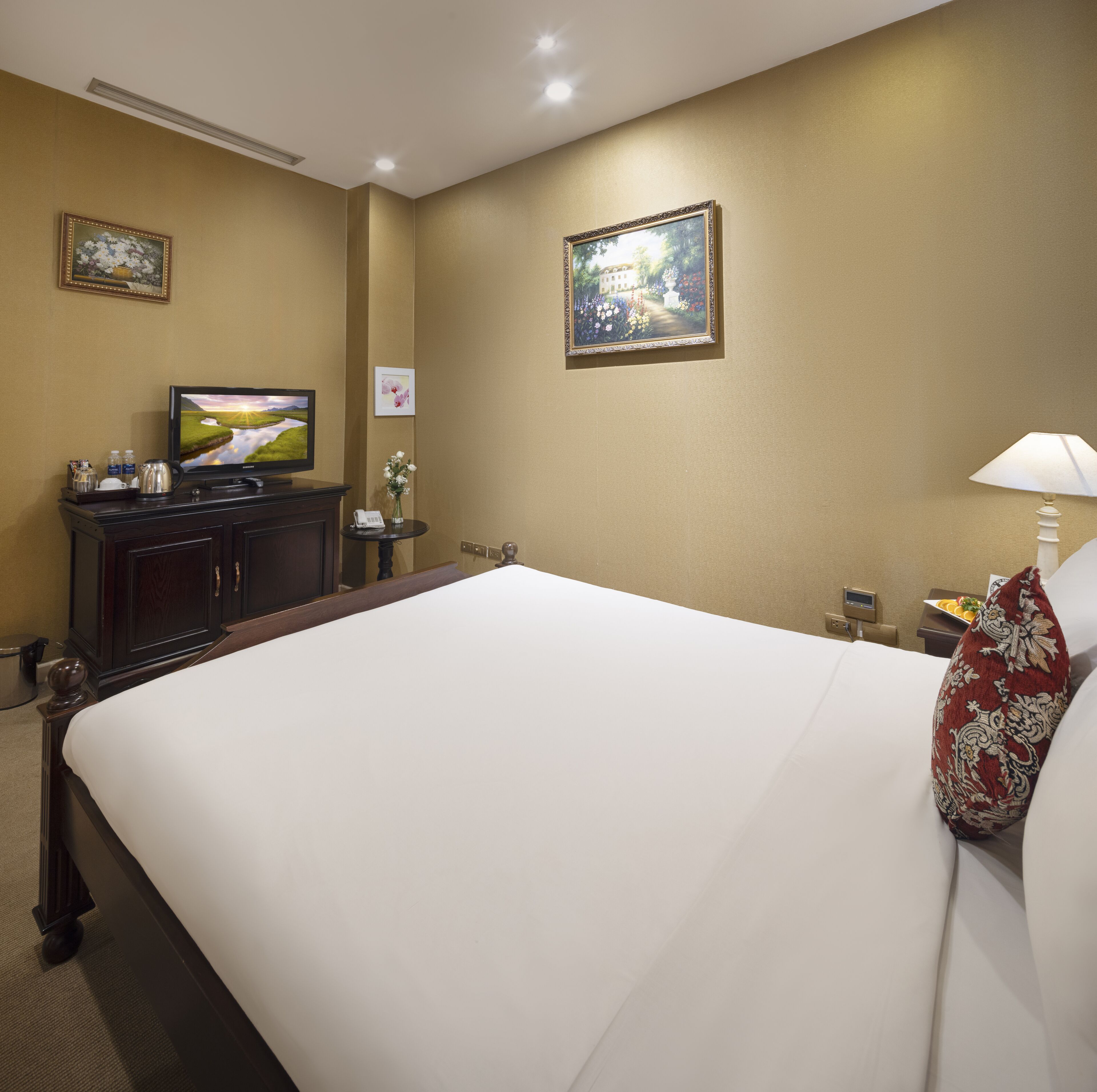 Une chambre d’hôtel avec un grand lit, un téléviseur posé sur une commode, une petite table avec un vase de fleurs et un tableau accroché au mur.