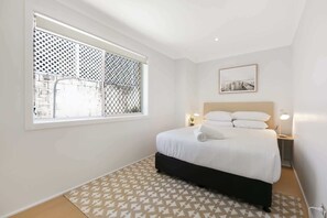 Une chambre à coucher avec un lit, une fenêtre ornée d’un treillis, une table de chevet avec une lampe et un tableau encadré accroché au mur.