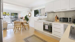 Une cuisine moderne avec des meubles blancs, un coin repas avec des chaises en osier et une vue à travers une porte vitrée.