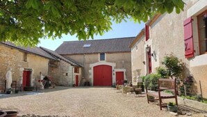 Une cour dallée de gravier, entourée de bâtiments en pierre aux portes et volets rouges.