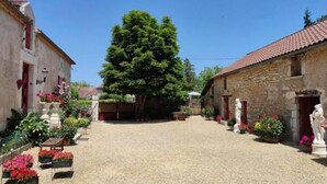 Une cour avec un grand arbre, des bâtiments en pierre et des parterres de fleurs.