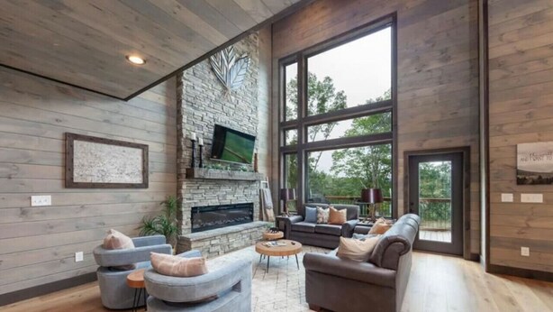 A spacious living room with a stone fireplace, large windows, and modern furniture. - Take A Dip Lodge - Indoor Heated Pool (Sevierville)