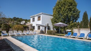 Ein Schwimmbecken mit Liegestühlen und Sonnenschirmen, ein weißes Gebäude mit Balkon und umgebende Grünflächen.