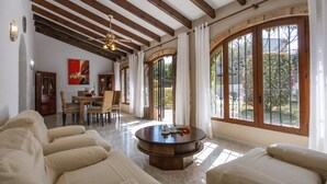 Ein geräumiges Wohnzimmer mit rundem Couchtisch, beigefarbenen Sofas, Holzbalken an der Decke und großen Fenstern mit Vorhängen.