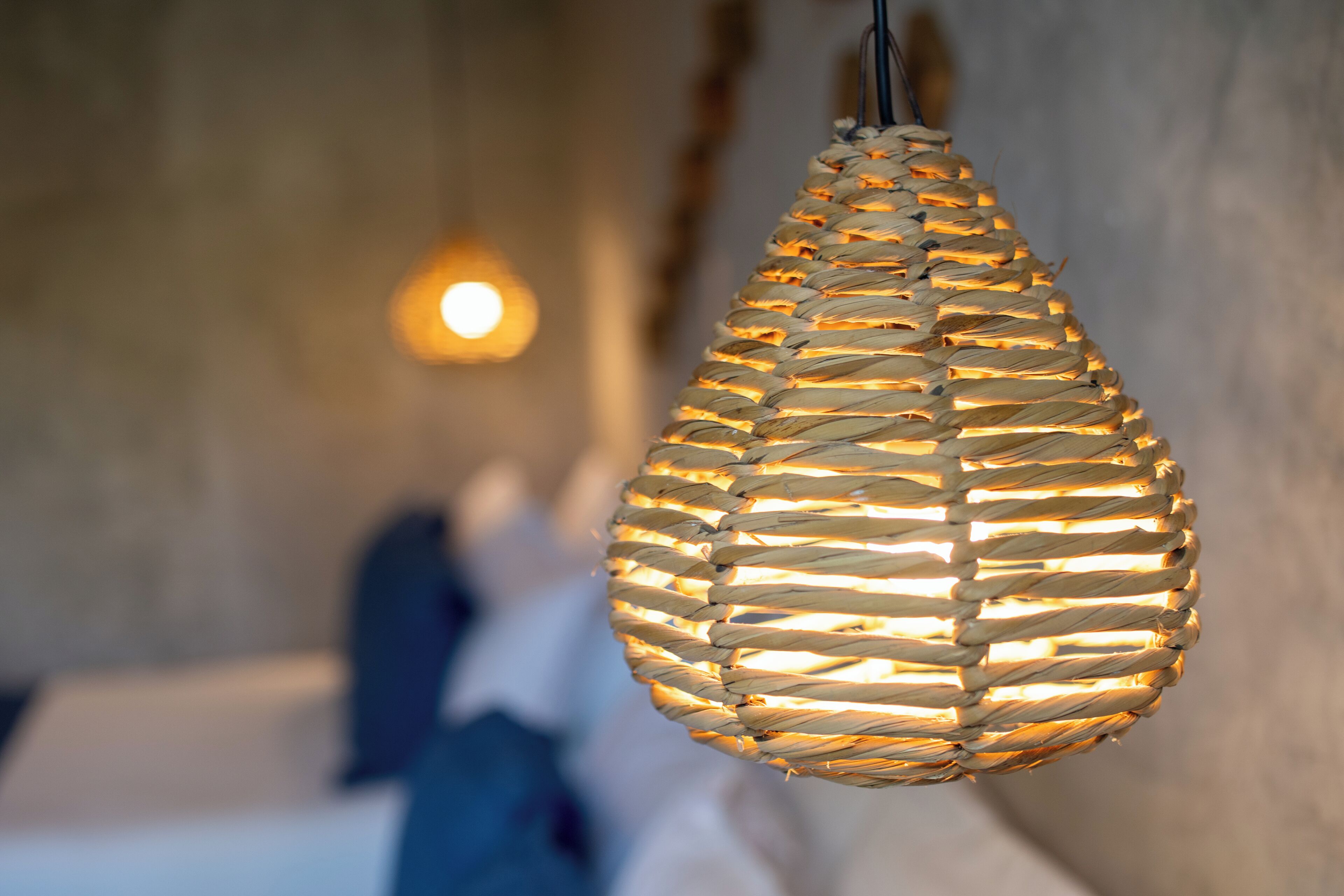 A woven light fixture hanging from the ceiling in a room with a neutral-colored wall and a bed with white bedding.