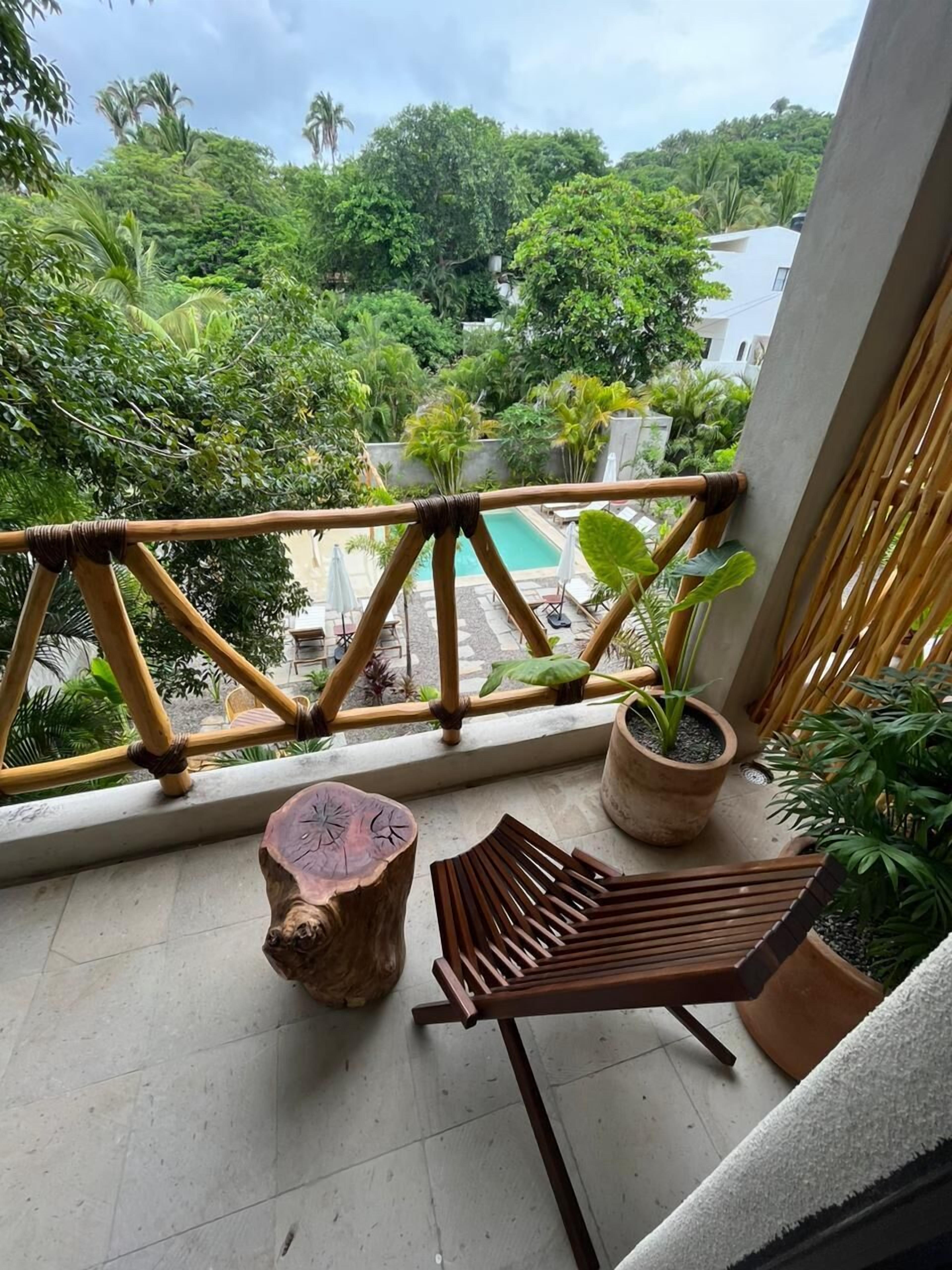 Un balcón con vista a una piscina, rodeado de exuberante vegetación y un edificio blanco al fondo.