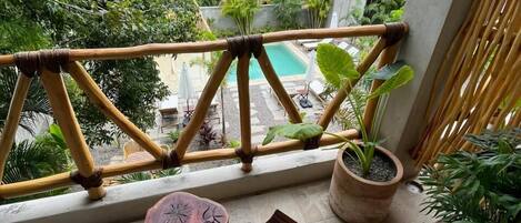 Un balcón con vista a una piscina, rodeado de exuberante vegetación y un edificio blanco al fondo.