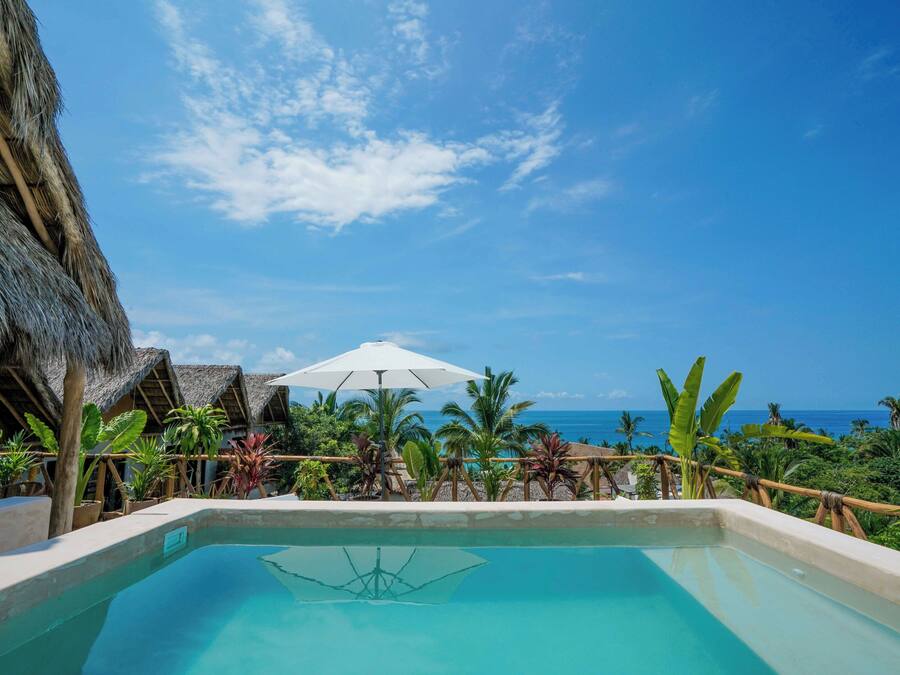 A pool area with a view of the ocean, surrounded by tropical plants and thatched-roof structures.