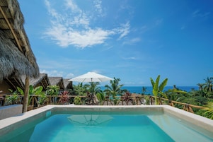 A pool area with a view of the ocean, surrounded by tropical plants and thatched-roof structures.