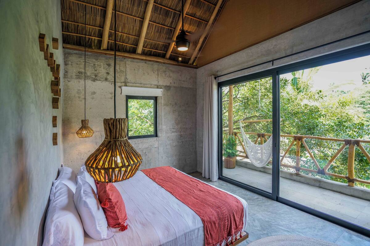 Un dormitorio con un ventanal grande, una cama con ropa de cama blanca y roja, una lámpara colgante y vistas de vegetación al exterior.