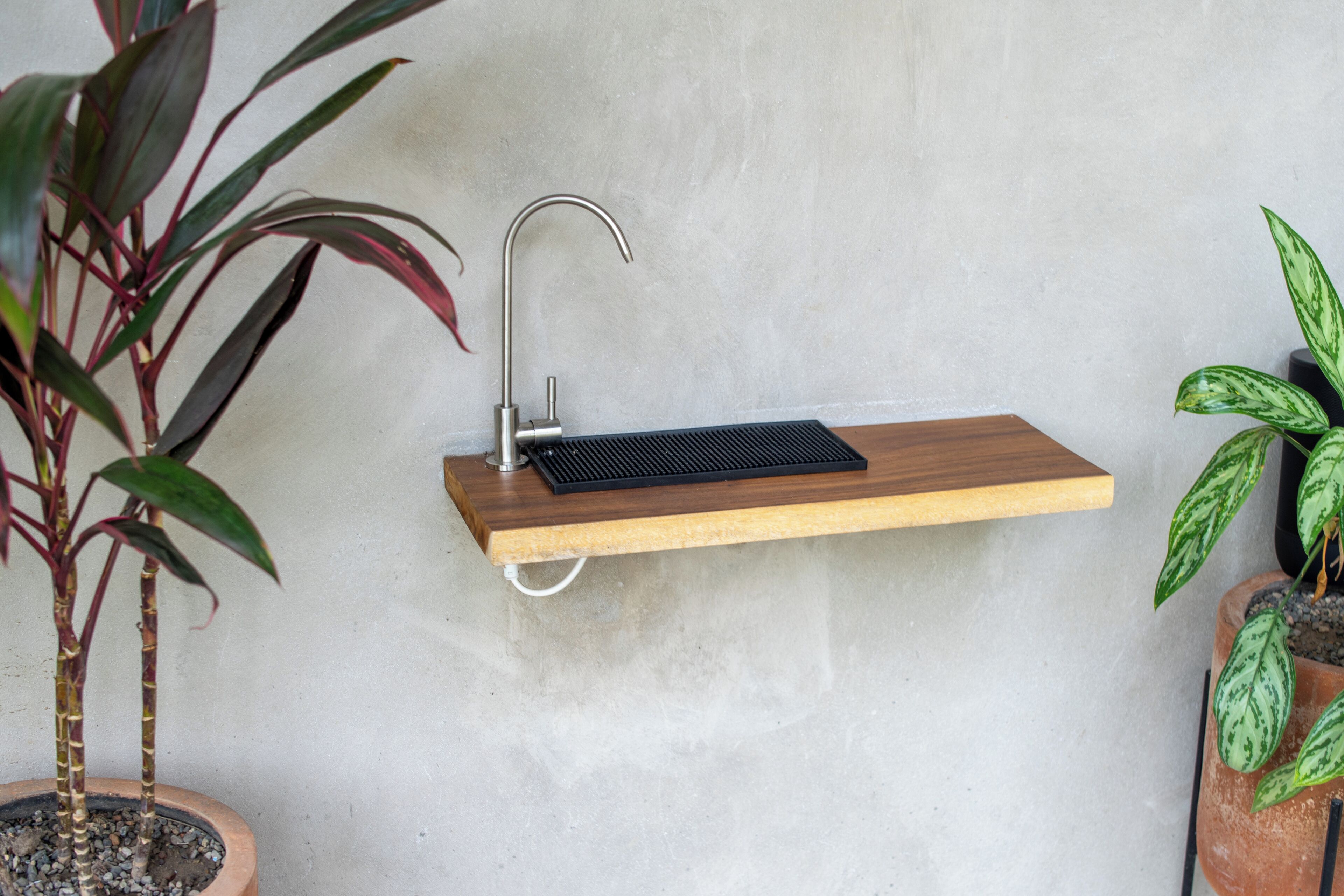 A wall-mounted sink with a wooden shelf, a metal faucet, and two potted plants.