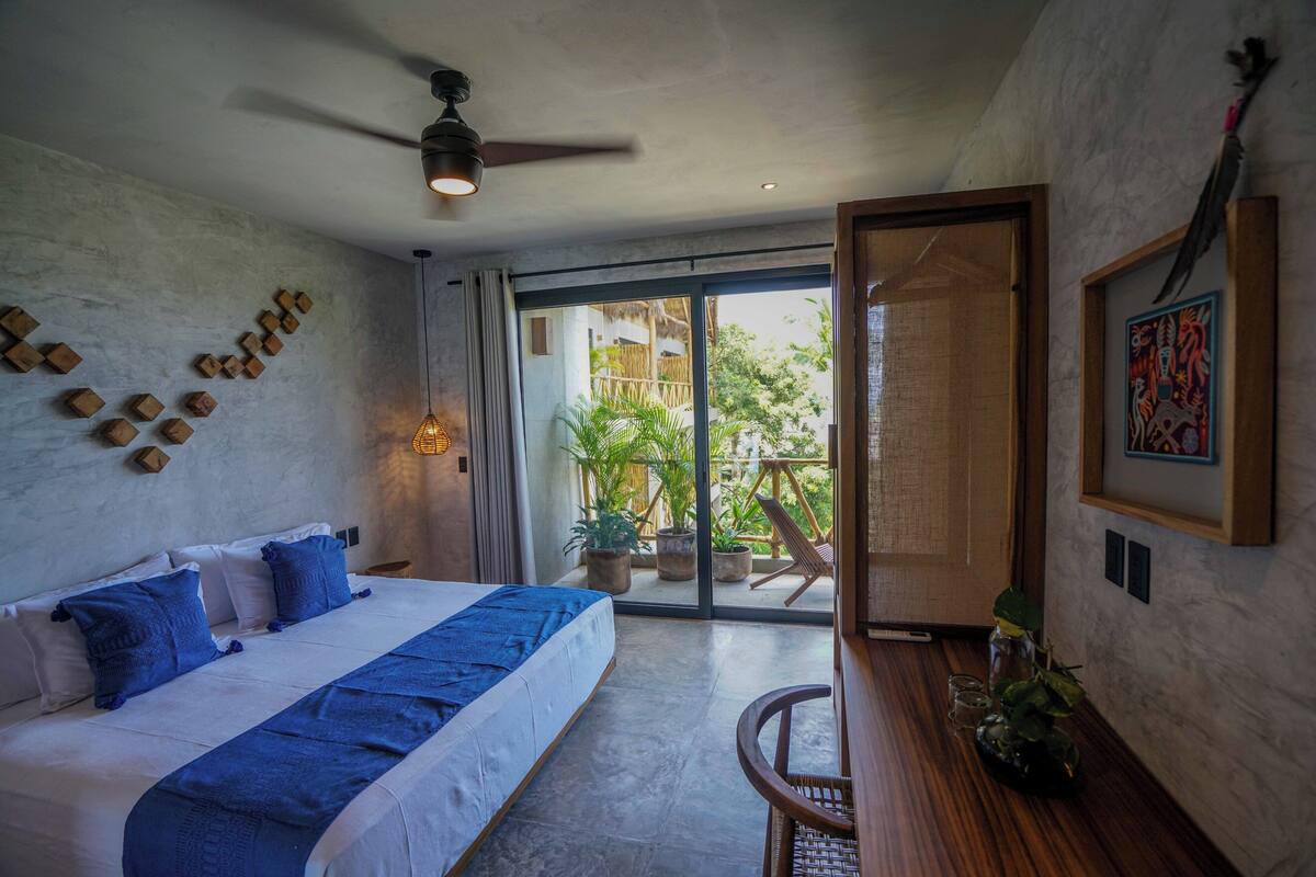 Un dormitorio con cama, ventilador de techo, vista al exterior, una mesa de madera con planta y una puerta que da a un balcón.