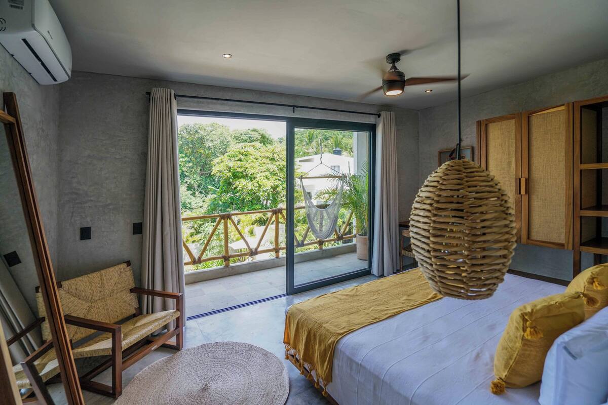 Un dormitorio con un gran ventanal corredera que da a un balcón, una cama con una colcha amarilla, una silla de madera y una lámpara colgante de ratán.