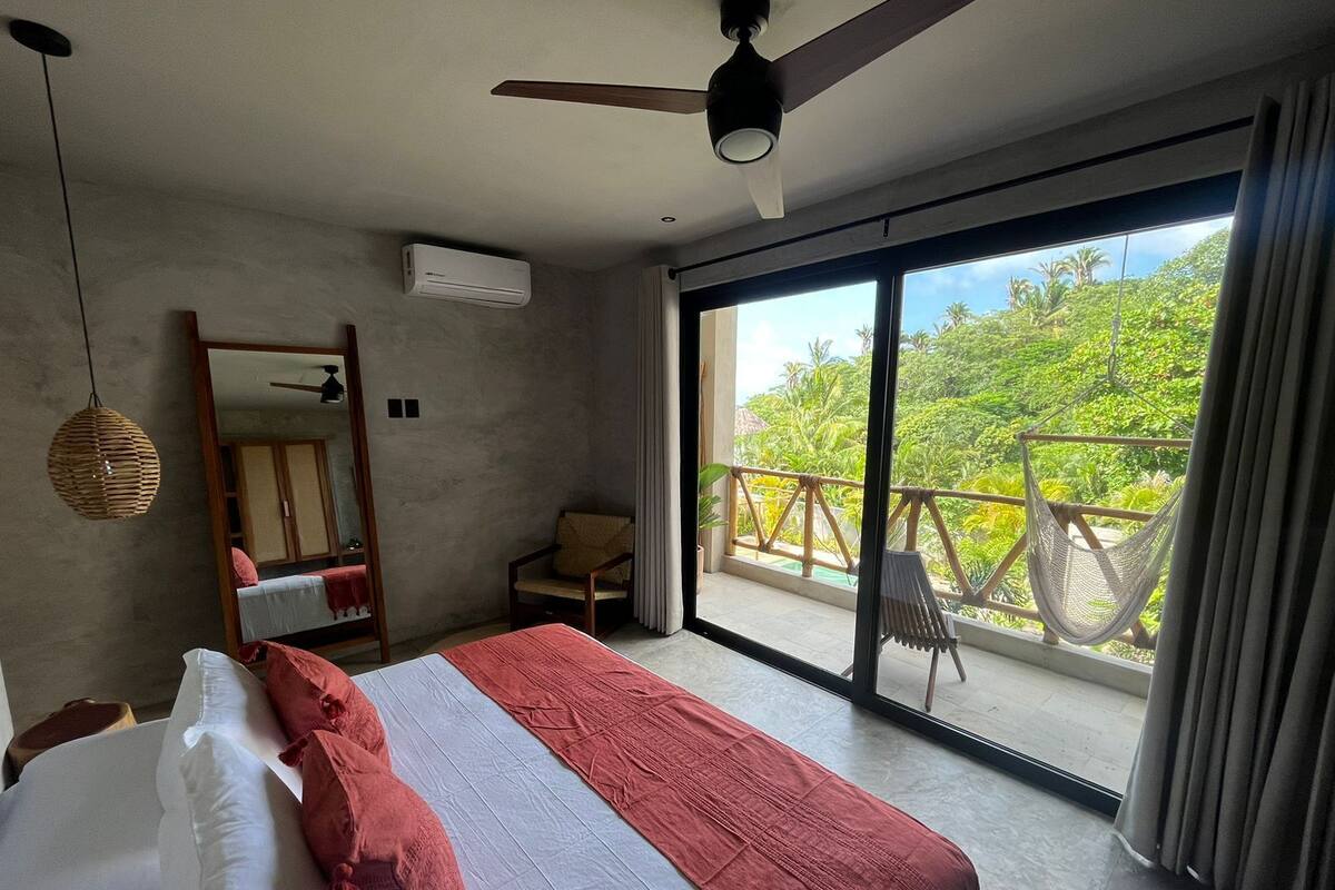 Un dormitorio con cama, ventilador de techo, equipo de aire acondicionado, una silla y un balcón con vistas a la vegetación.
