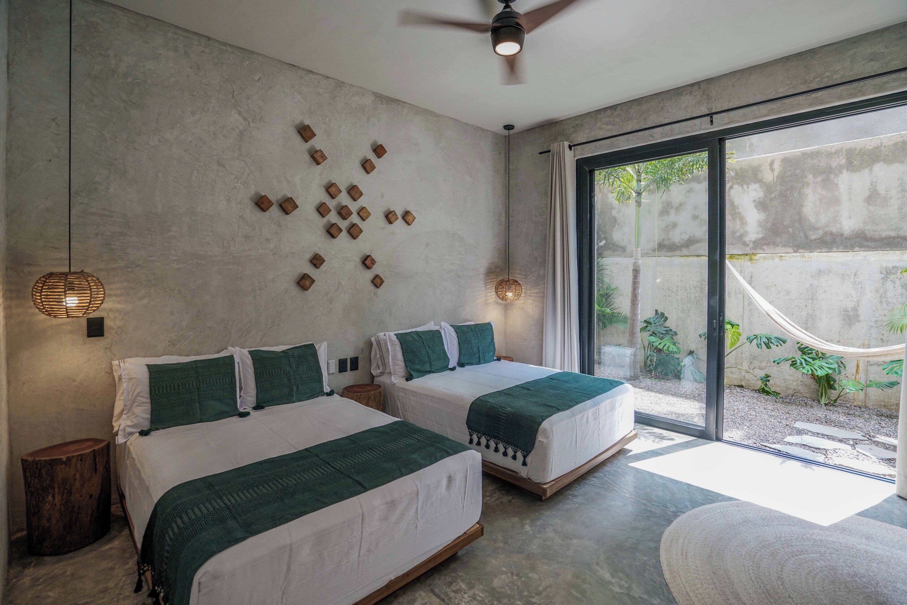 A modern bedroom with two beds, a concrete wall, a ceiling fan, and a large sliding glass door leading to a garden.