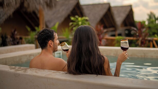 A couple relaxing in a hot tub with drinks, surrounded by thatched-roof huts.