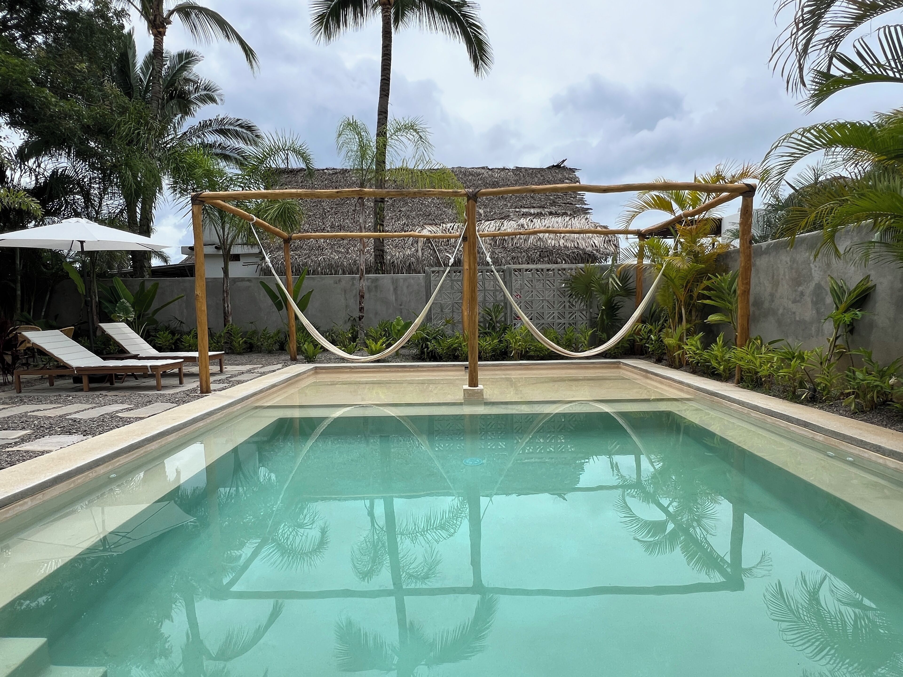 Ein Pool mit einer Holzpergola und einer Hängematte, umgeben von Palmen und Liegestühlen.