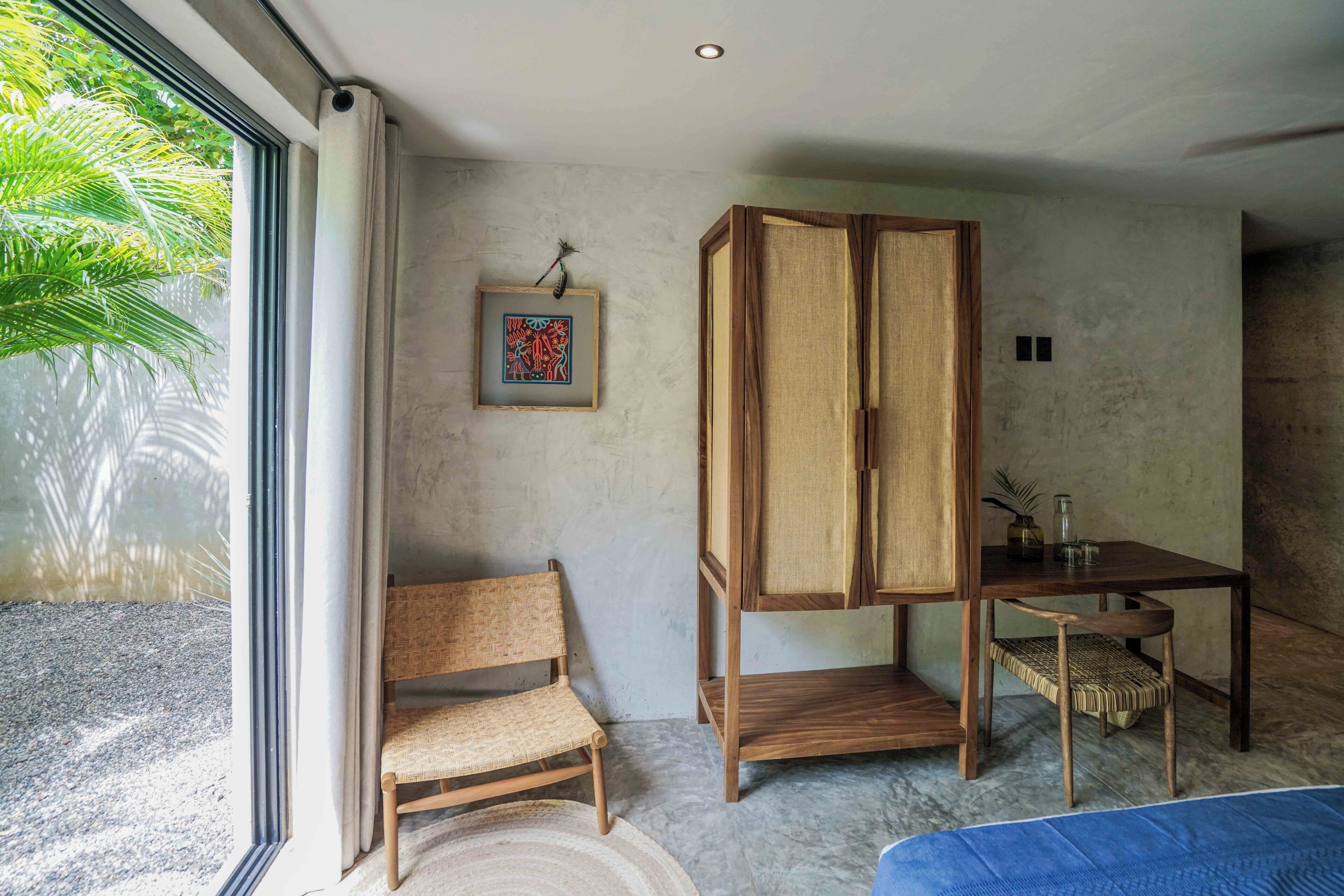 Een kamer met een groot raam, een houten stoel, een houten kast, een klein tafeltje met een vaas en een lijst met een foto aan de muur.