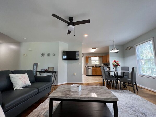 A modern living room with a black leather sofa, a marble coffee table, and a dining area with a table and chairs. A kitchen is visible in the background with wooden cabinets and a window.